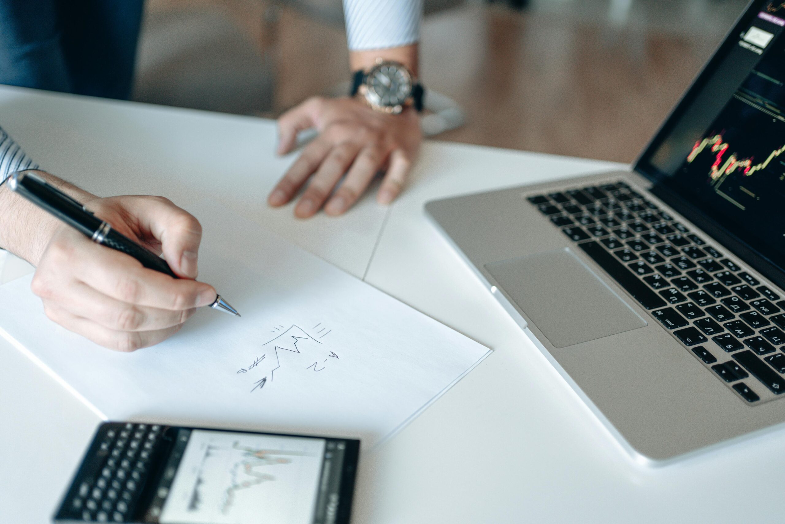 Close-up of hands drawing stock trends on paper with laptop and smartphone on desk.